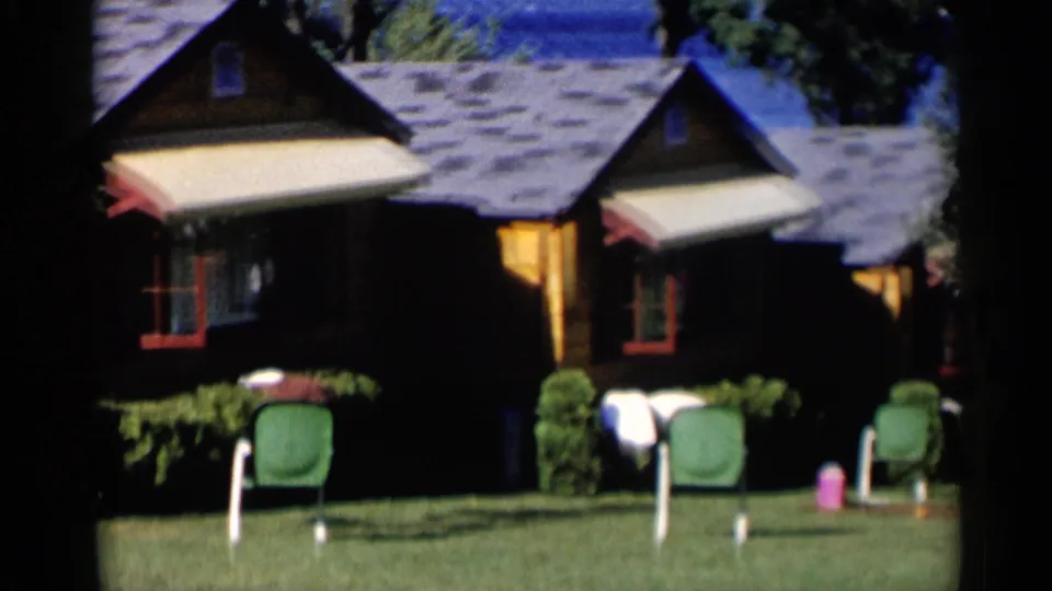 Lake George trip footage cuts to a small building or cabin with awnings, lawn chairs, shrubs, and bright 1950s home-movie color.