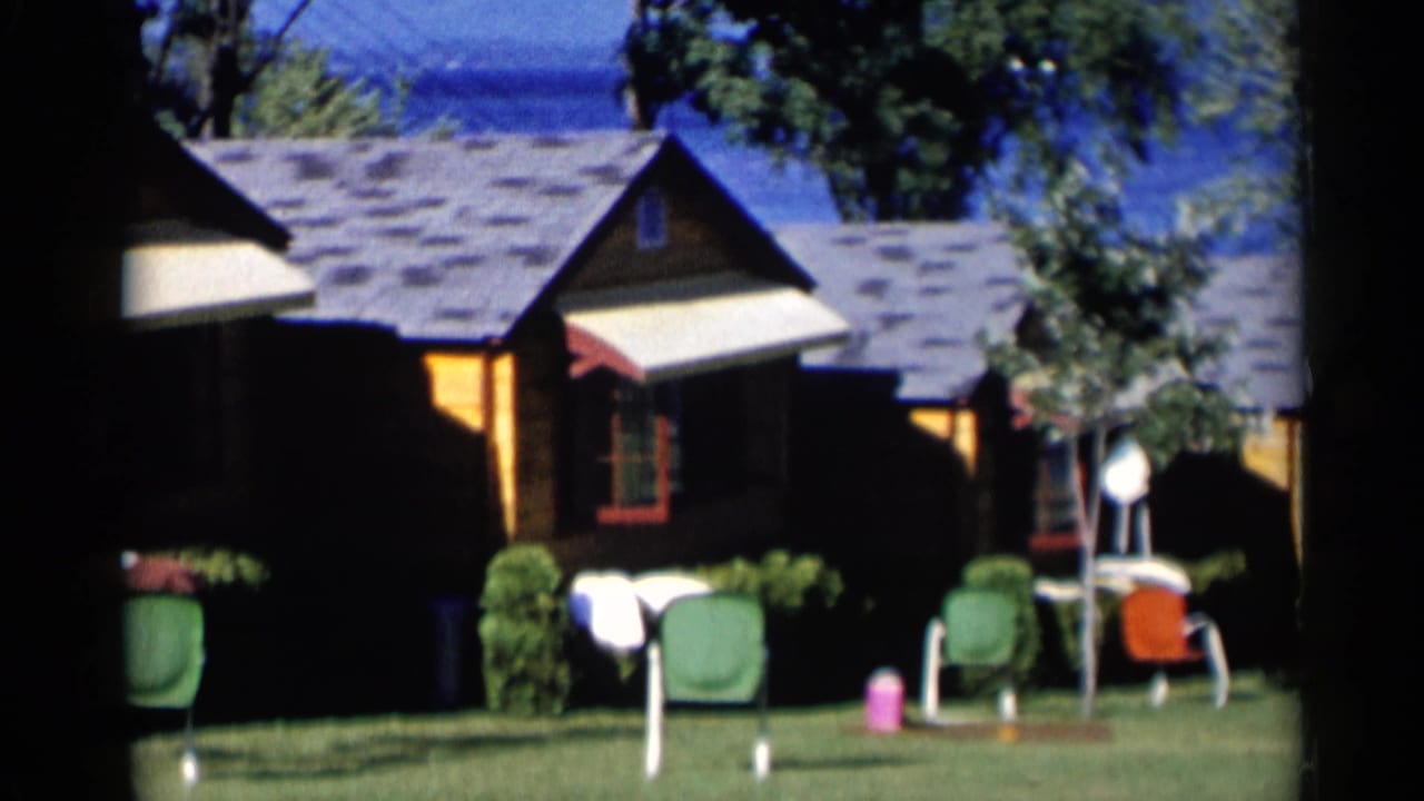 1955 Lake George home-movie frame showing rustic cabins, green lawn chairs, shrubs, and blue lake water beyond the roofs.