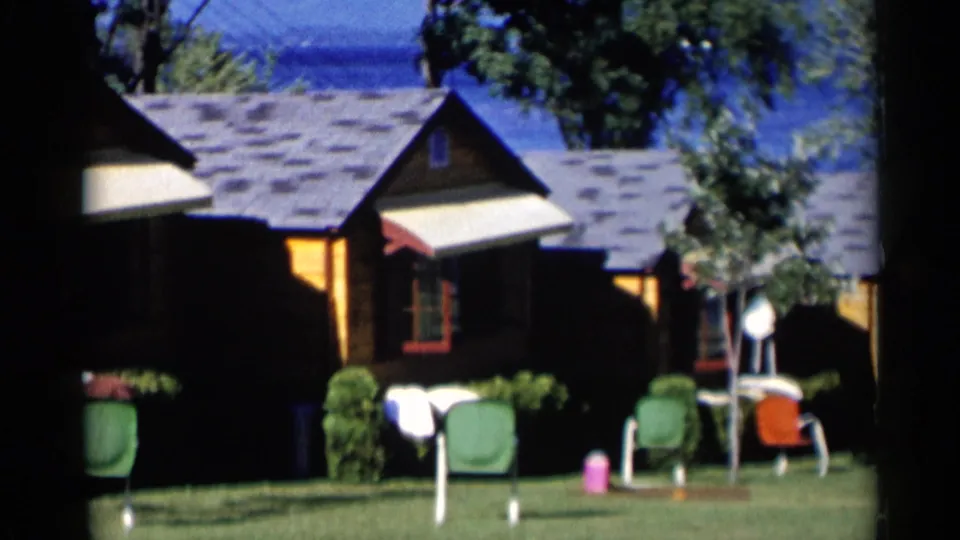 1955 Lake George home-movie frame showing rustic cabins, green lawn chairs, shrubs, and blue lake water beyond the roofs.