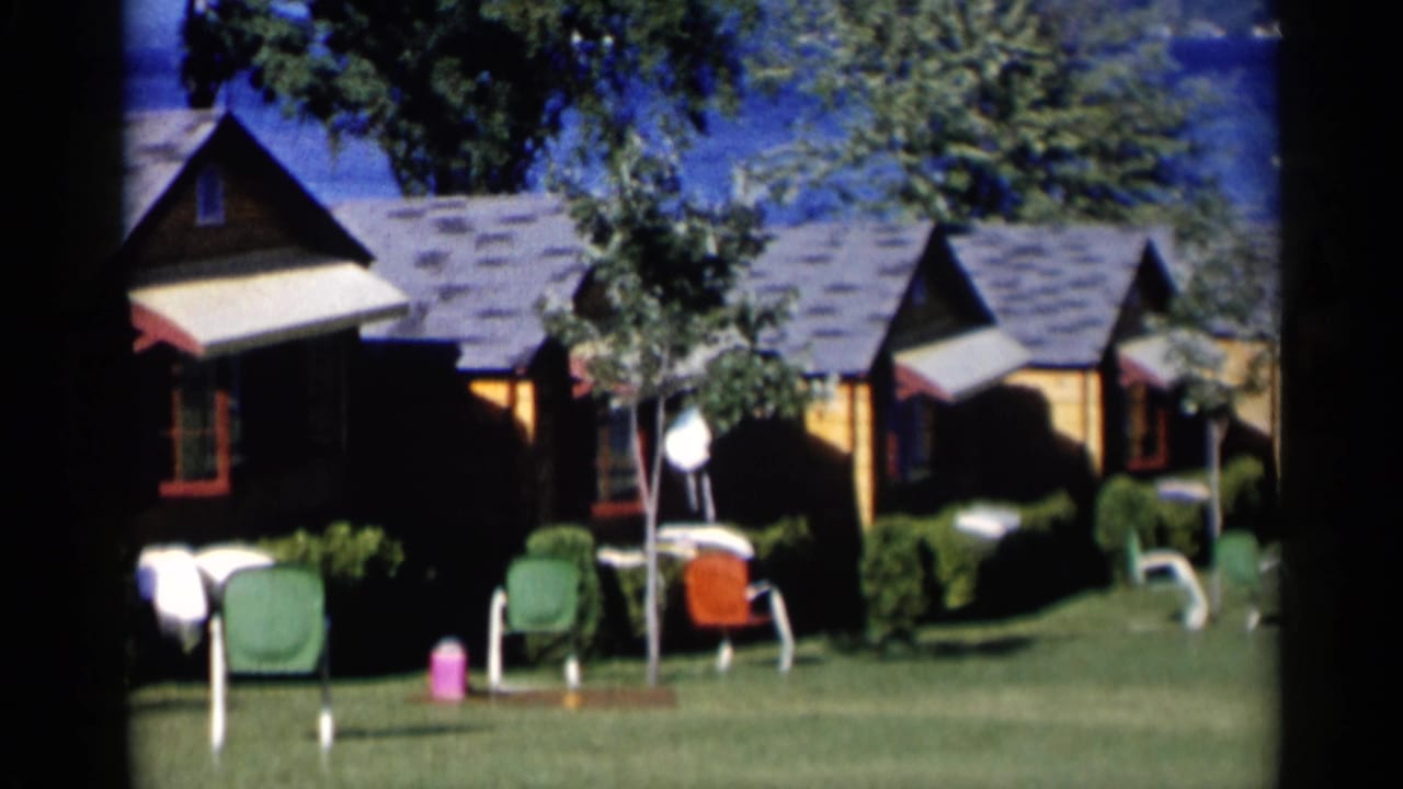 Lake George vacation cabins with striped awnings, green and orange lawn chairs, trimmed shrubs, and lake water in the background.