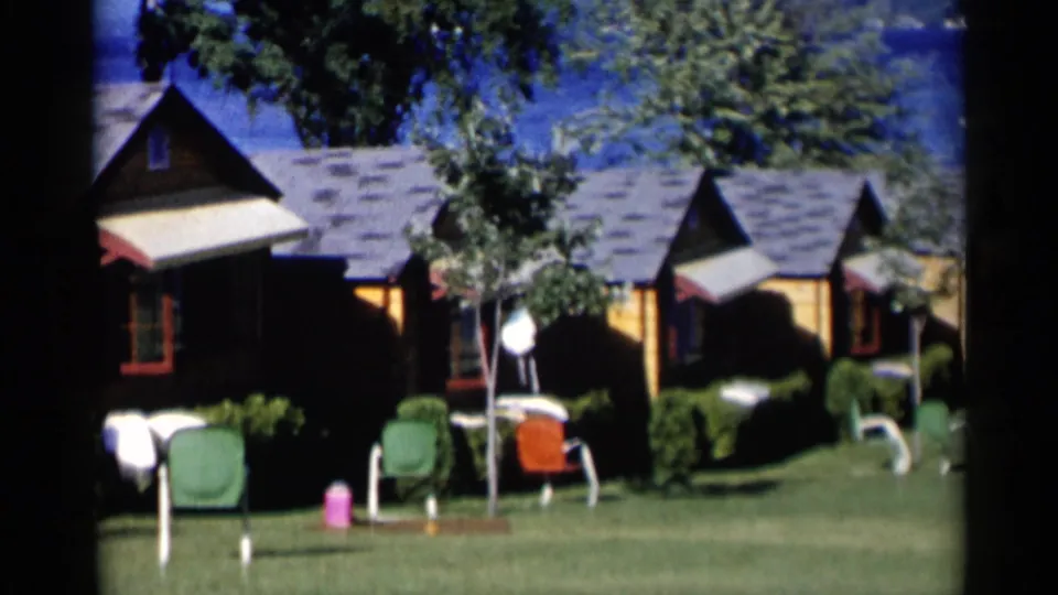 Lake George vacation cabins with striped awnings, green and orange lawn chairs, trimmed shrubs, and lake water in the background.