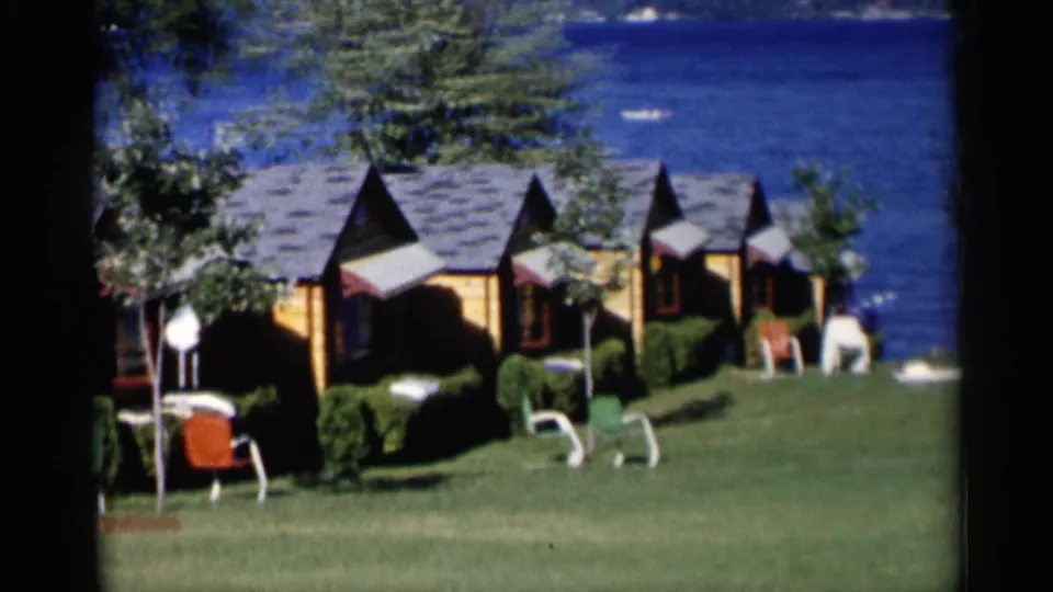1950s Lake George frame showing a row of wooden vacation cabins, scattered lawn chairs, open grass, and blue lake behind them.