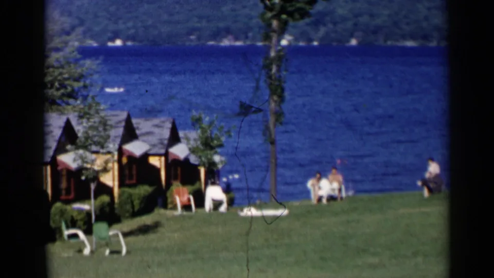 Lake George home-movie frame with cabins on the left, open grass, blue lake water, and several people seated near the shoreline.
