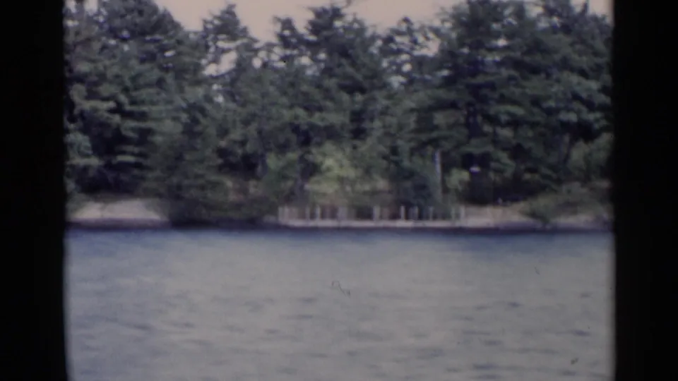 1955 home-movie view across Lake George toward dense shoreline trees and a small pier partly obscured by foliage.