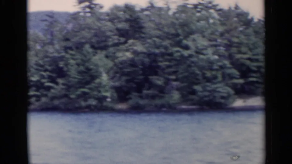 1955 8mm view of Lake George with calm water, tree-covered shore, and a forested mountain in the distance.