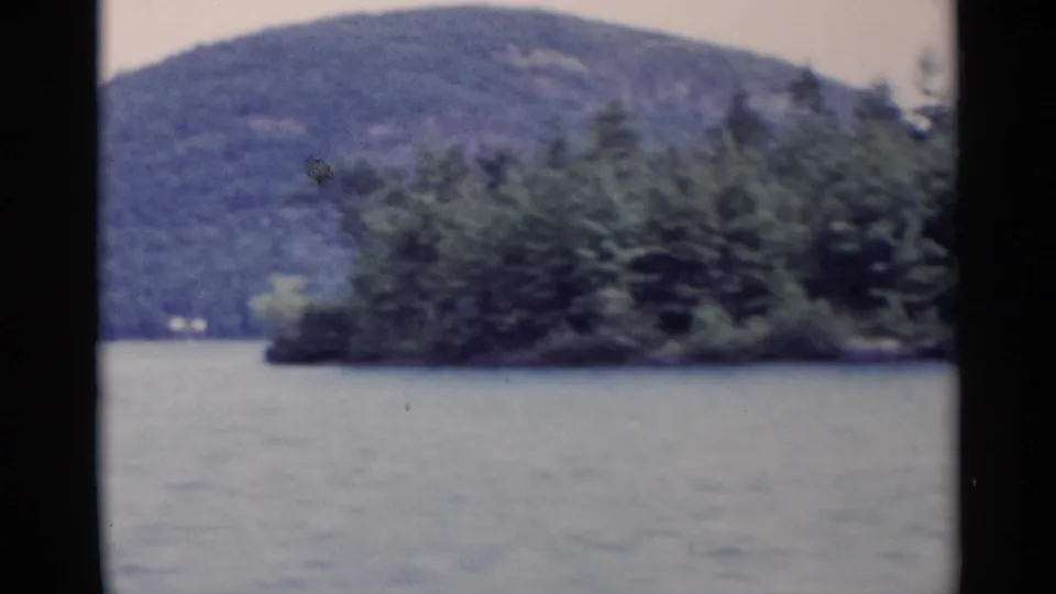 1955 Lake George home-movie frame showing open water, a tree-covered island, and a hazy mountain backdrop.
