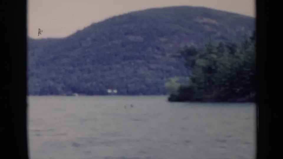 1955 8mm Lake George view with rippled water, forested hillside, and a dark wooded point along the shore.