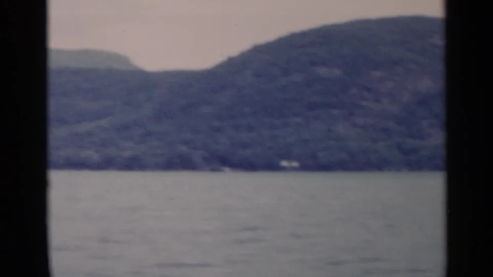 1955 Lake George 8mm frame showing a wide stretch of water, layered forested hills, and a small wooded shoreline at right.
