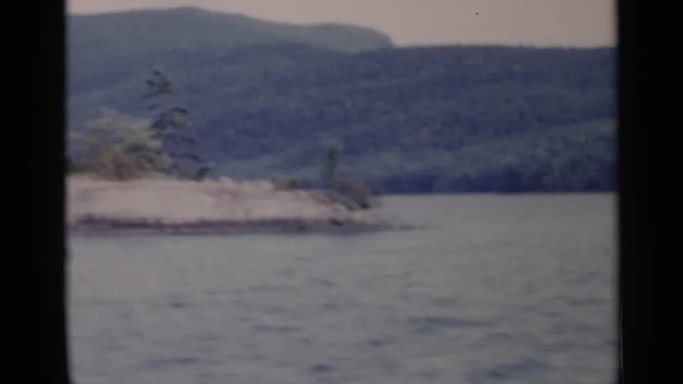 1955 Lake George view of a low rocky point or small island with evergreens and wooded hills behind it.