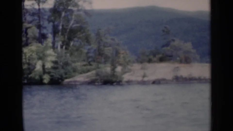 1955 Lake George frame showing a wooded shoreline, pale rocky ground, and distant hills across calm water.