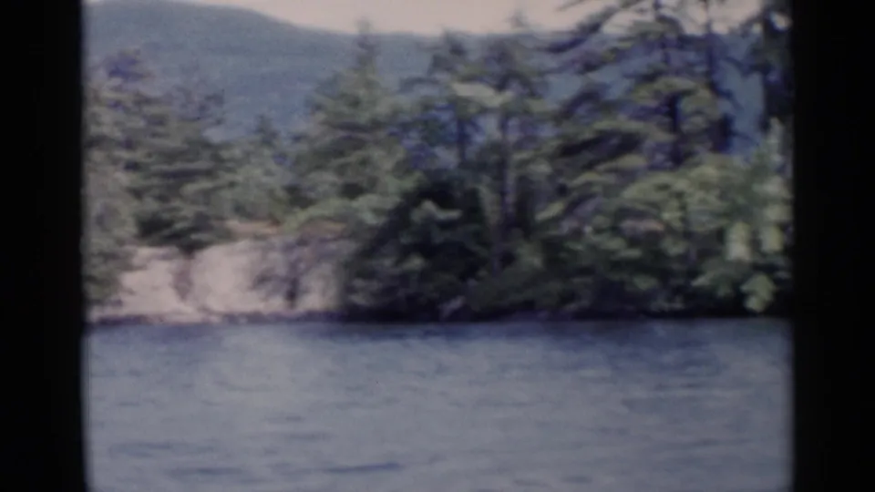 1955 Lake George home-movie frame with dense evergreens on a rocky shoreline and hills beyond the water.