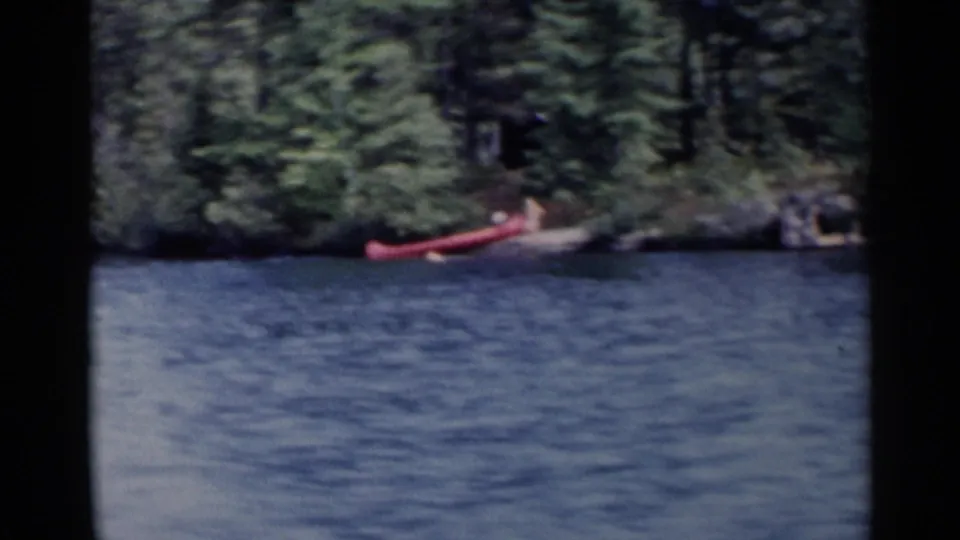 1955 Lake George 8mm frame showing a red canoe near a wooded shoreline across calm blue water.