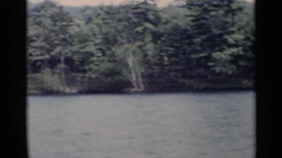 Lake George shoreline in a 1955 8mm home-movie frame, with rippled water and dense green trees.