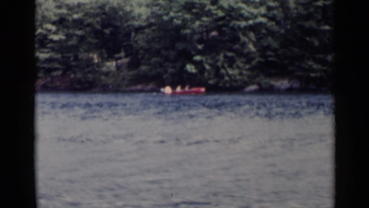 A red canoe crosses Lake George, New York, with dense wooded shoreline behind it in 1955 8mm home-movie footage.