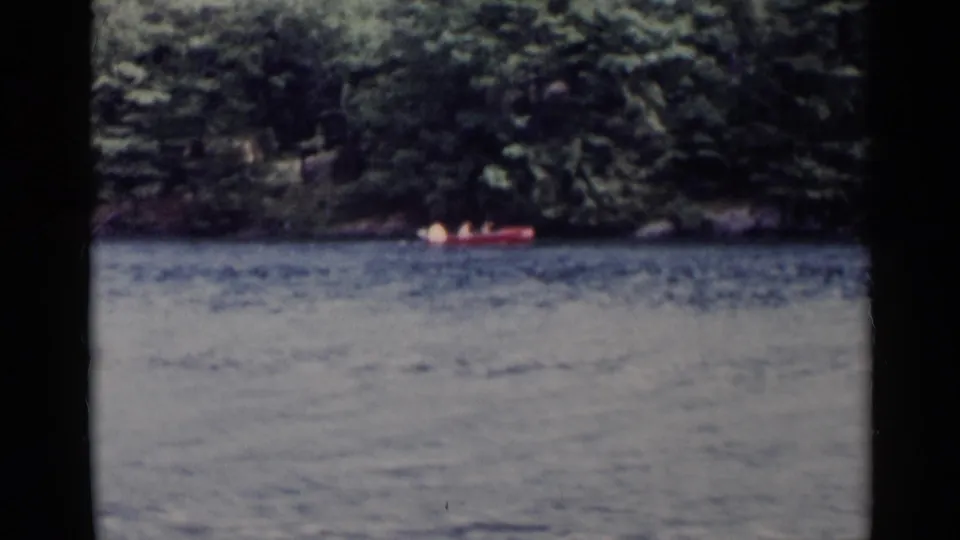 A red canoe crosses Lake George, New York, with dense wooded shoreline behind it in 1955 8mm home-movie footage.