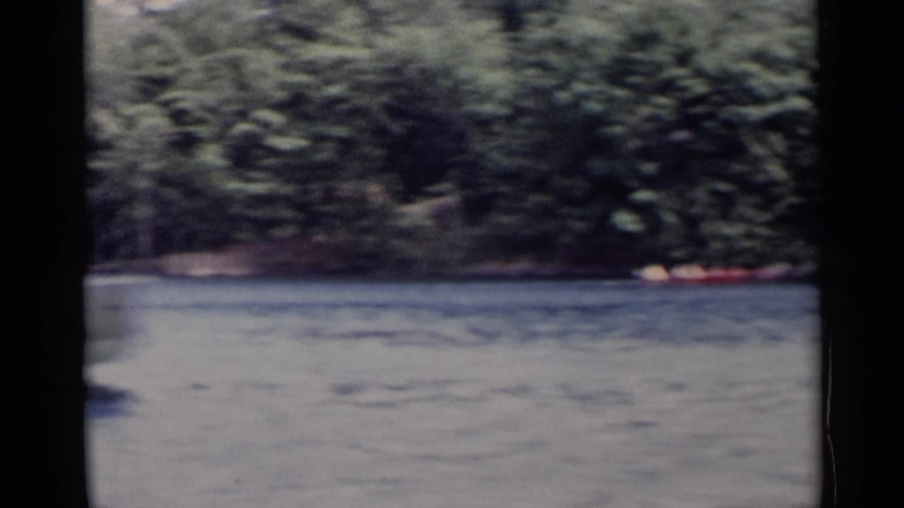 Distant boaters in a red canoe travel near the tree-lined shore of Lake George in mid-1950s home-movie footage.