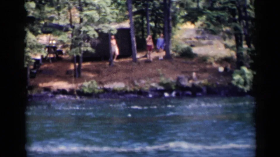 People gather along the Lake George shoreline near a rustic lakeside cabin area, seen from across the water in 1955 8mm footage.