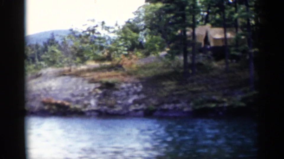1955 Lake George frame with a small rustic cabin or shelter partly hidden by trees above the rocky shoreline.