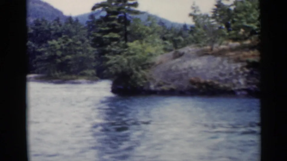 Lake George home-movie view of a rocky tree-covered point with hills beyond and calm blue water in front.