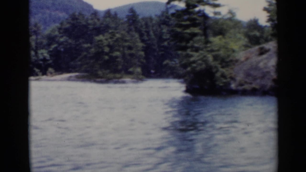 1955 Lake George home-movie frame showing rippled water, a wooded shoreline, distant hills, and dark film gate edges.