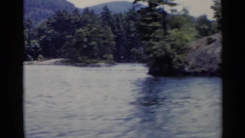 1955 Lake George home-movie frame showing rippled water, a wooded shoreline, distant hills, and dark film gate edges.