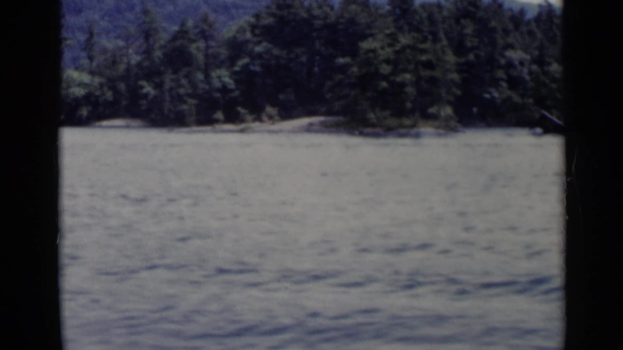 Lake George shoreline in a 1955 8mm frame, with open water, dense trees, low hills, and visible dark frame borders.