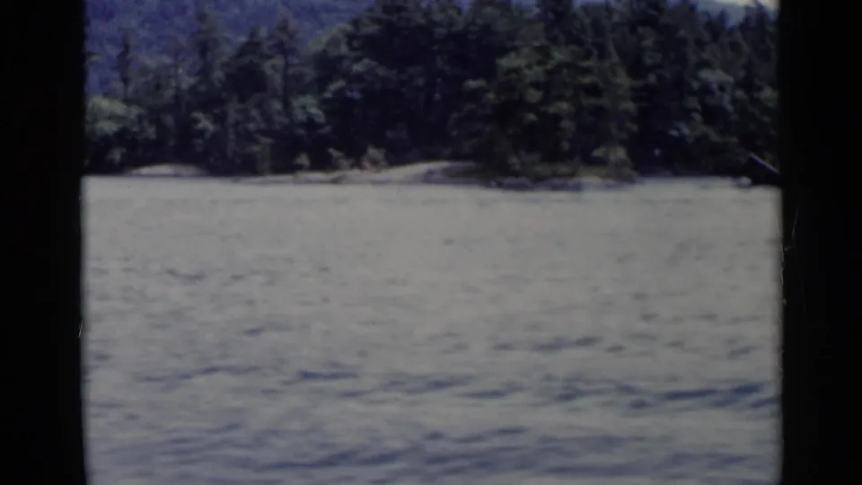 Lake George shoreline in a 1955 8mm frame, with open water, dense trees, low hills, and visible dark frame borders.