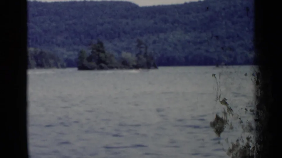 1955 Lake George view across rippled water toward a wooded hill and small island or shoreline point, with film wear visible near the frame edge.