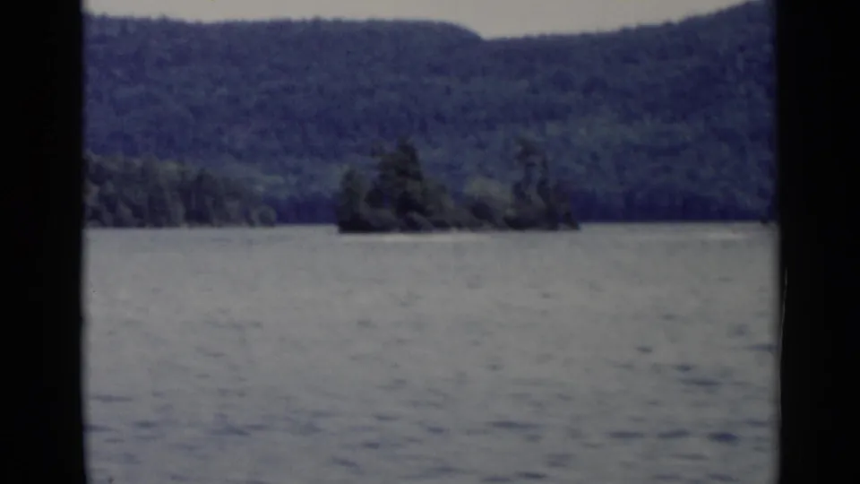 Lake George 1955 home-movie frame showing a small wooded island, rippled water, forested shore, and dark 8mm frame borders.