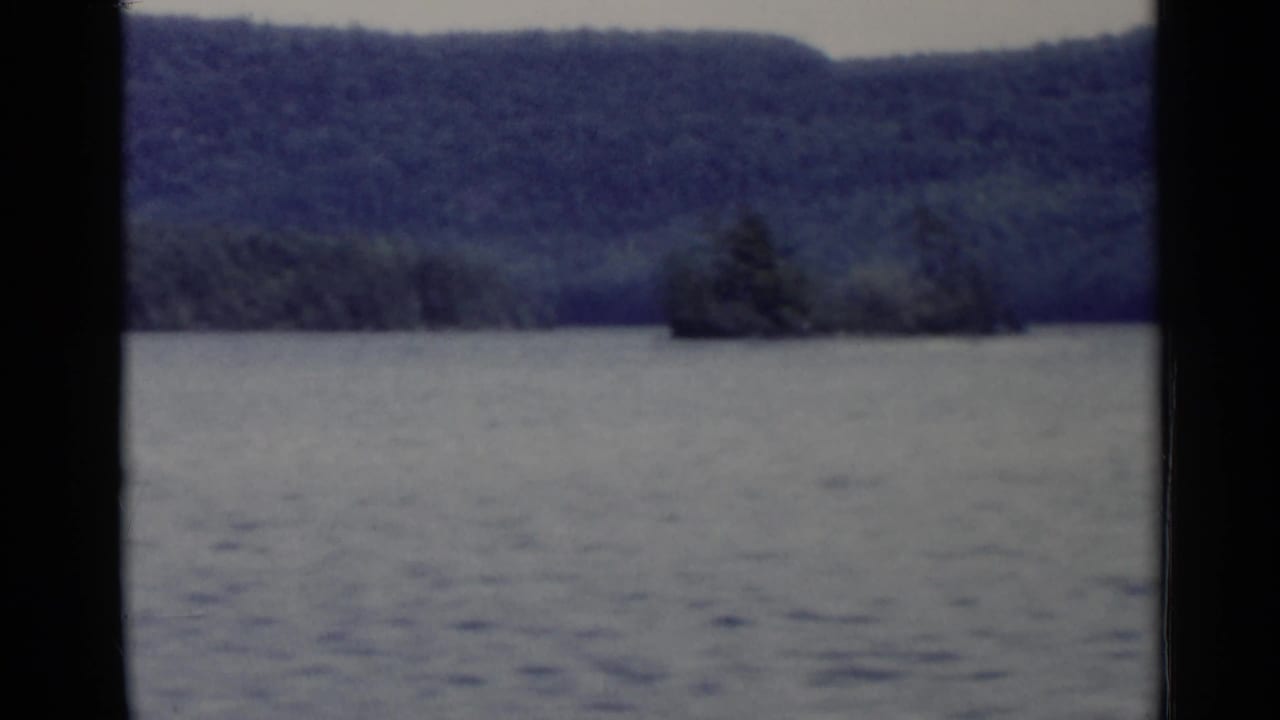 1955 Lake George 8mm view of open water with forested hills and a small tree-covered island in the distance.