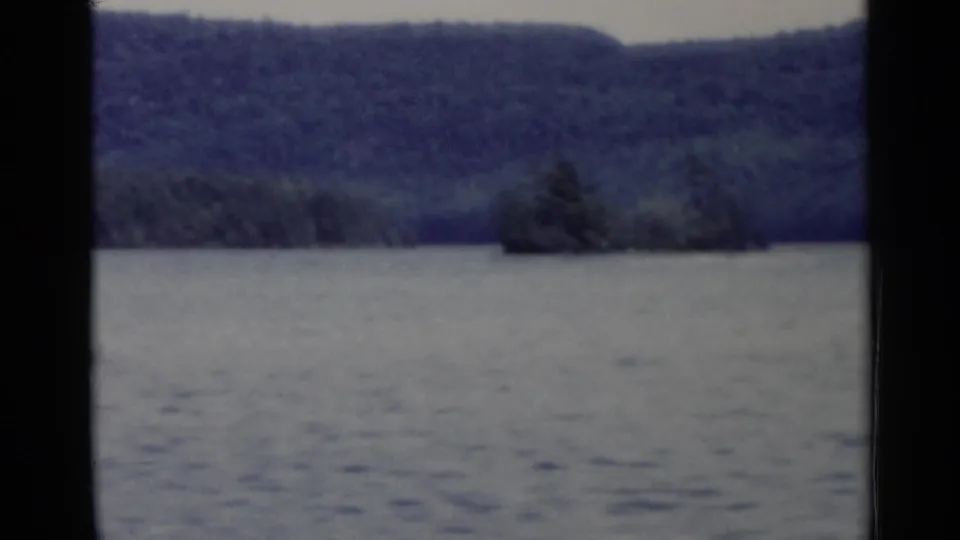 1955 Lake George 8mm view of open water with forested hills and a small tree-covered island in the distance.