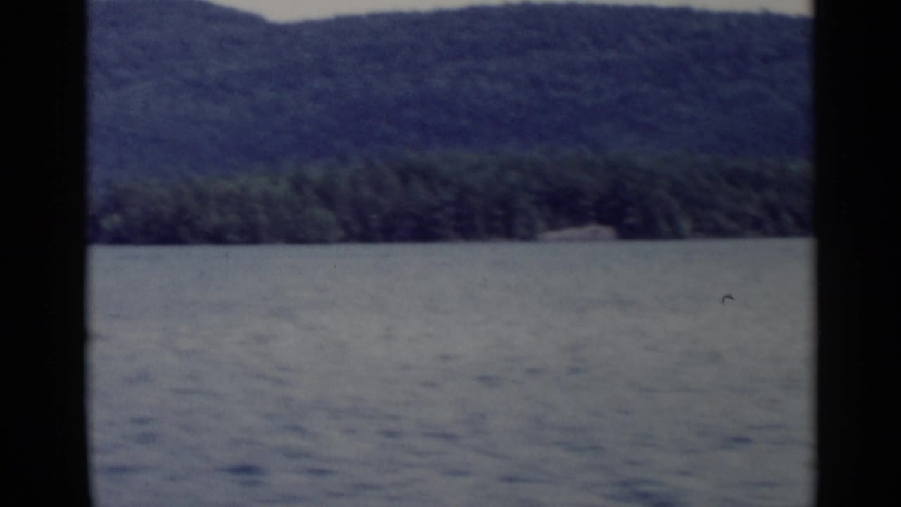 Lake George shoreline appears beyond rippled water, with wooded hills and soft 8mm color from the 1955 home movie.
