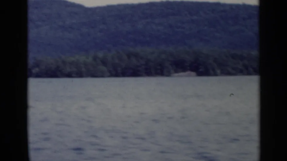 Lake George shoreline appears beyond rippled water, with wooded hills and soft 8mm color from the 1955 home movie.