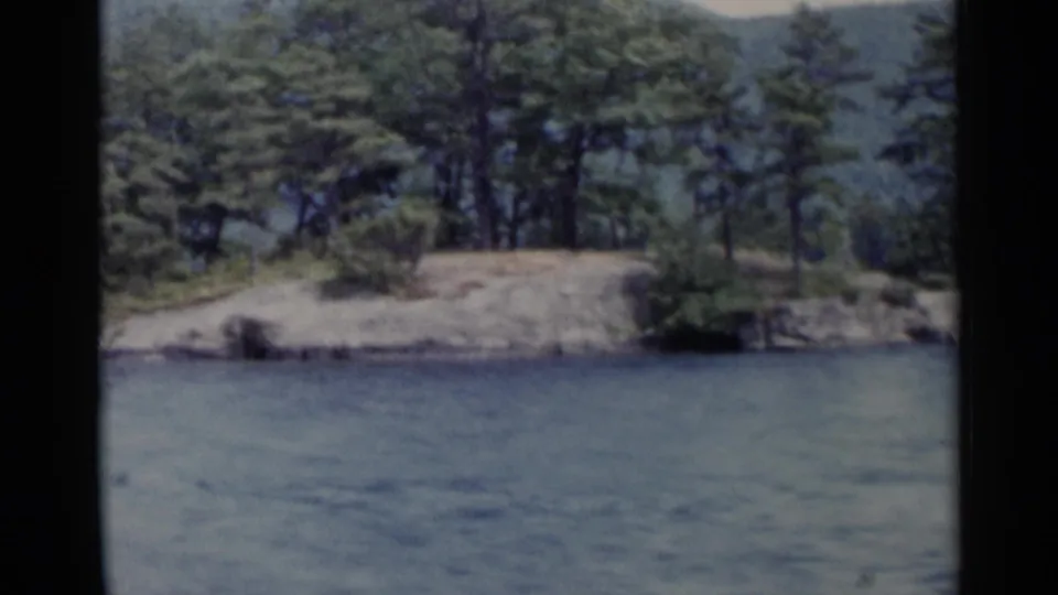 A tree-covered island with a rocky edge fills the frame as the Lake George boat passes in 1955 8mm footage.