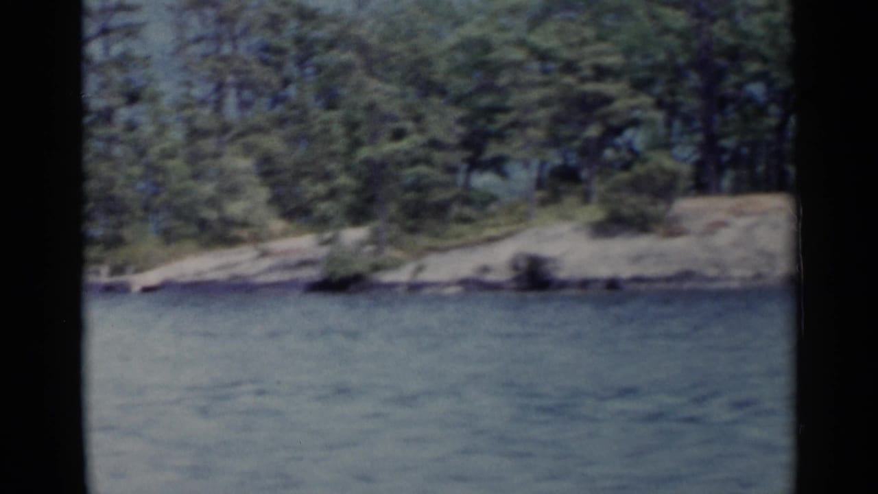 1955 Lake George 8mm frame showing calm water, rocky wooded shoreline, evergreen trees, and hazy hills beyond.