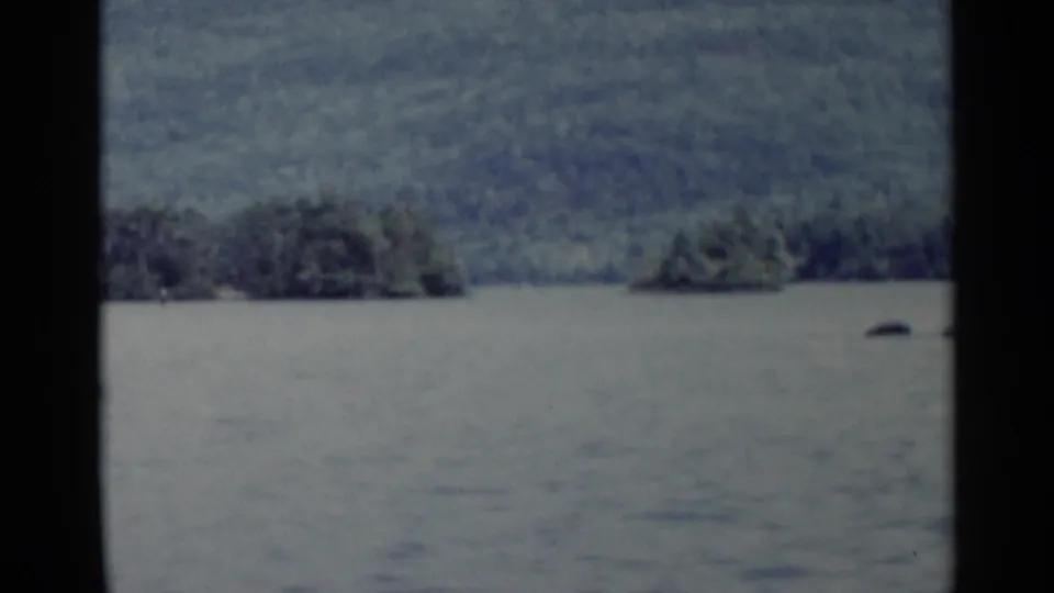 1955 Lake George footage with open water, small wooded islands, distant hills, and the vehicle window frame in view.