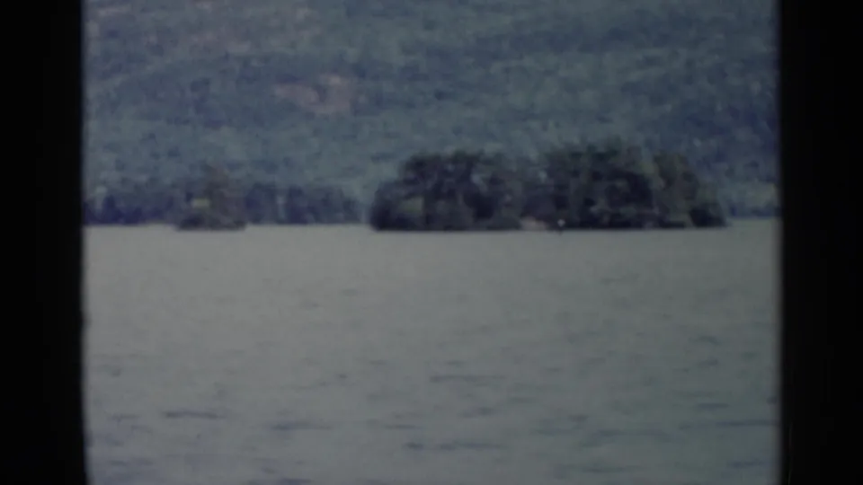 Lake George landscape with calm water, a low wooded island, distant forested hills, and soft 8mm film texture.
