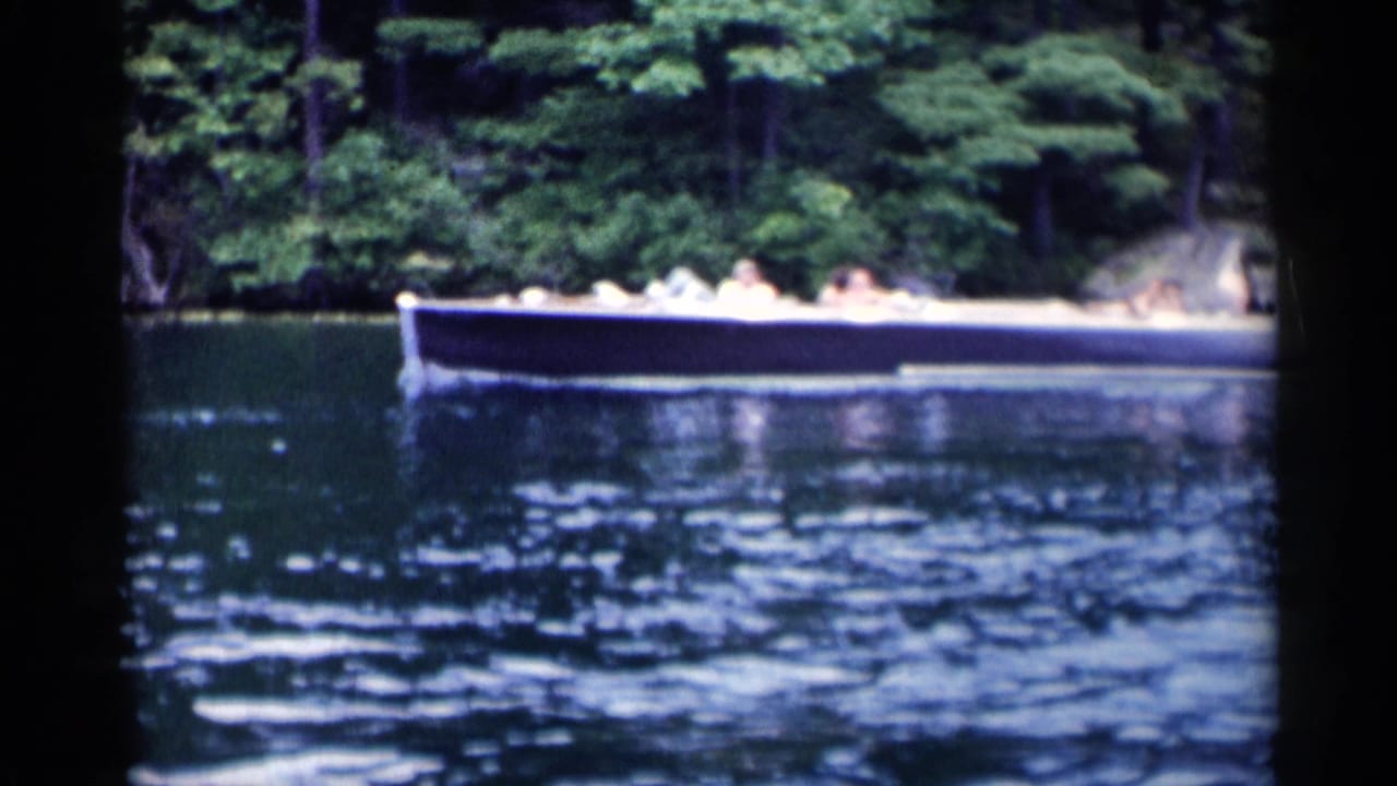 A low motorboat with distant passengers moves across Lake George near a dense wooded shoreline in 1955 8mm footage.