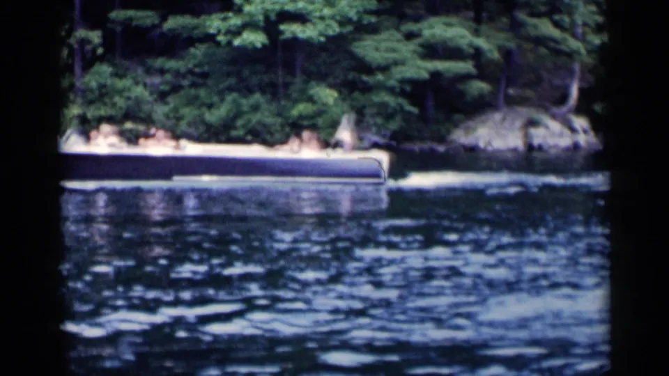 A passenger motorboat travels across rippled Lake George water, with trees and rocks behind it in vintage 1955 home-movie footage.