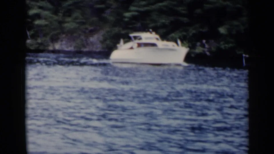 A white motor yacht cruises on Lake George near shaded trees and shoreline rocks in soft 1955 8mm home-movie footage.