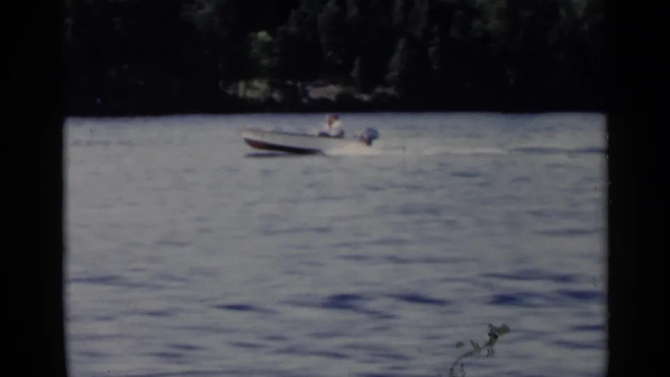 Small motorboat with a distant person aboard moves across Lake George in soft 1950s 8mm color.