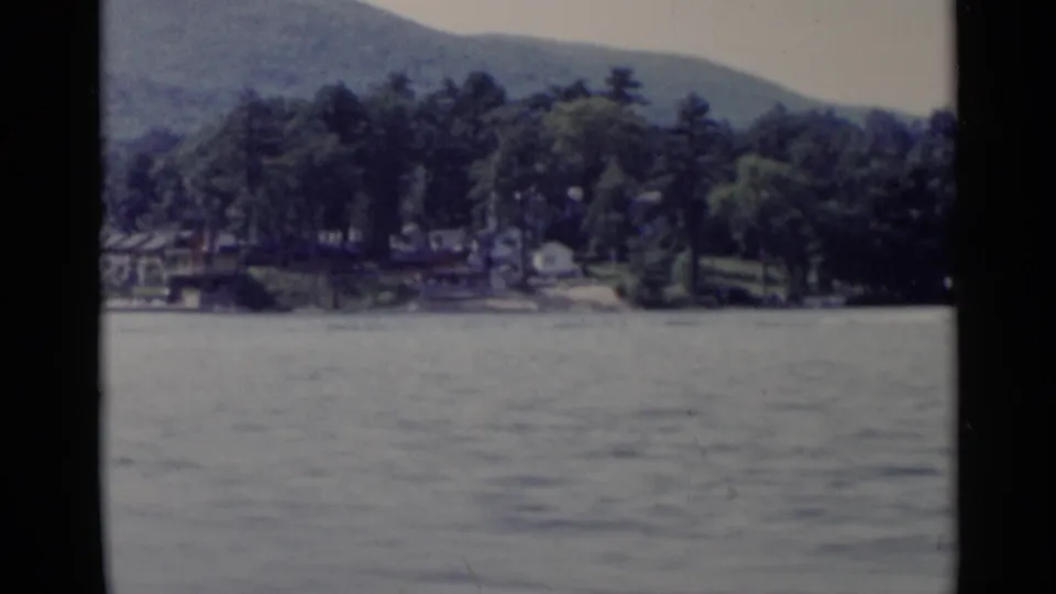 Lake George shoreline view with trees, distant lakeside structures, water, and hills in a 1955 home-movie frame.