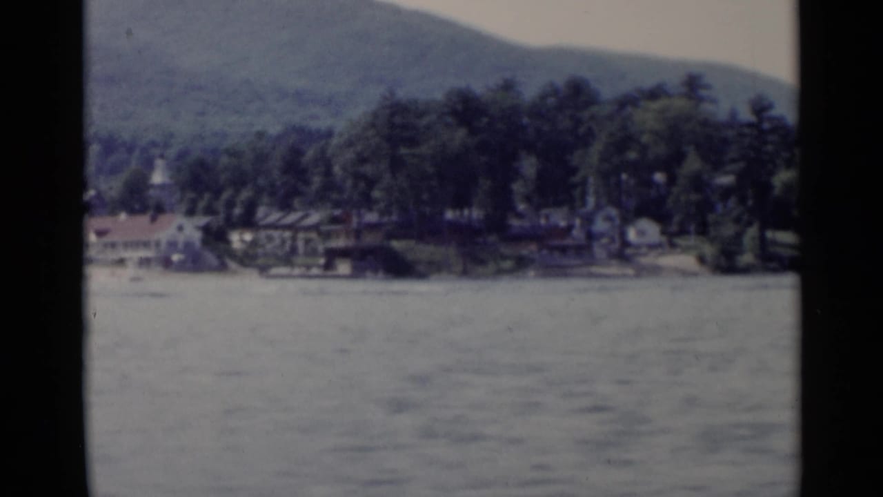 Lake water facing a wooded shoreline with low mountains beyond.