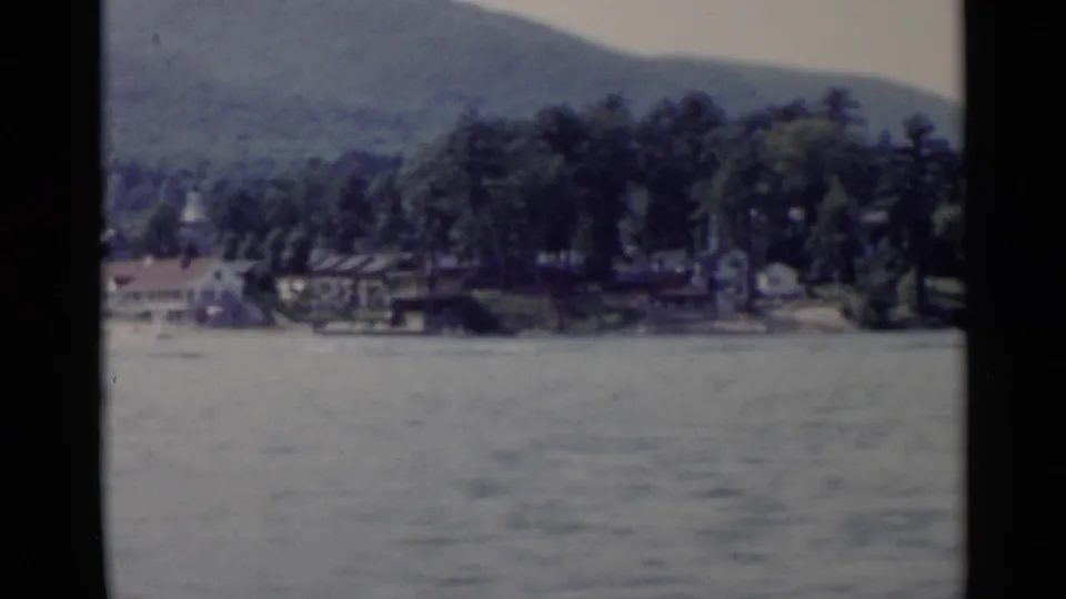 1955 Lake George view across calm water toward trees, lakeside buildings, and distant mountains.