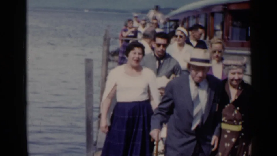 At Lake George in 1955, adults in suits, hats, dresses, and sunglasses walk along a pier beside the water.