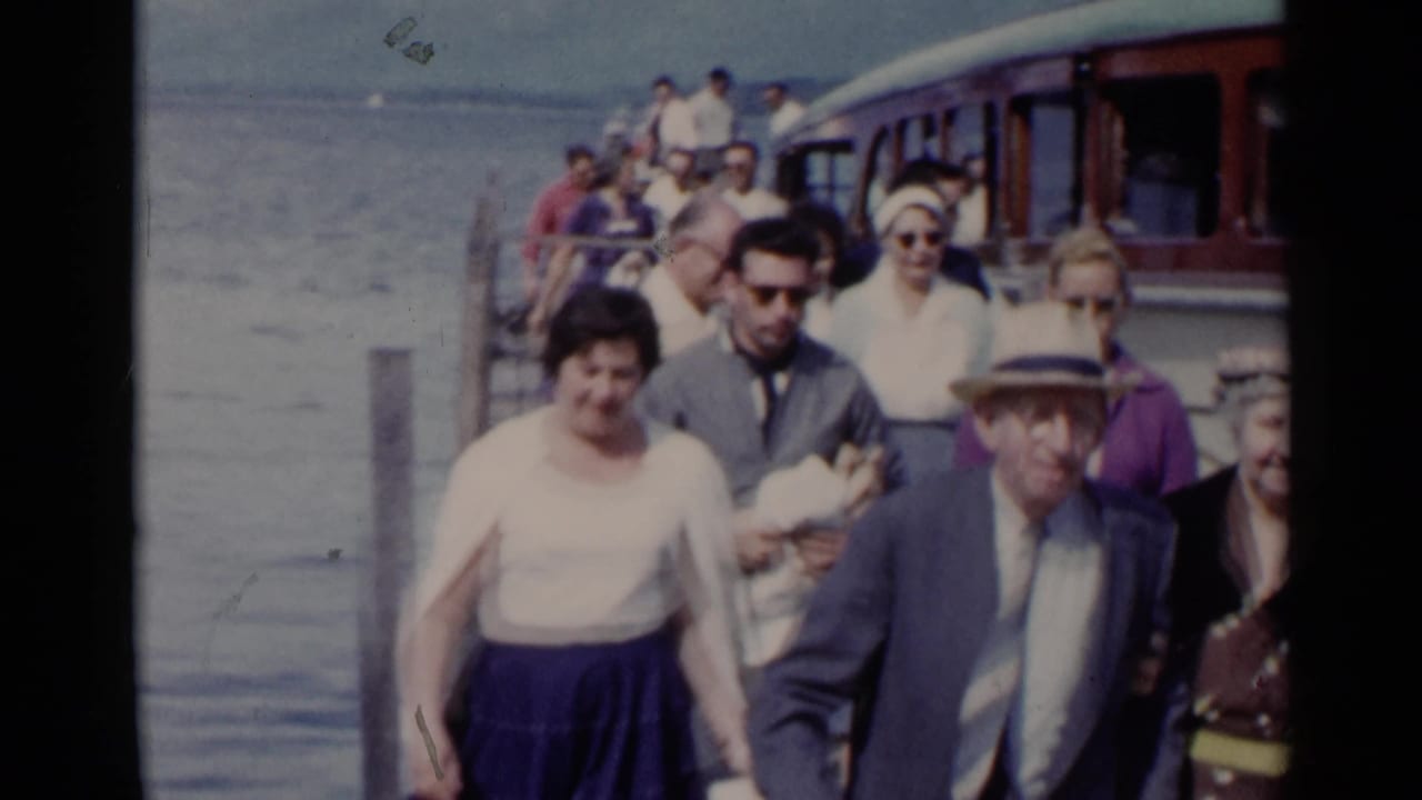 In Lake George, New York, 1955 home-movie footage, adults in mid-century clothing walk on a dock beside the lake.