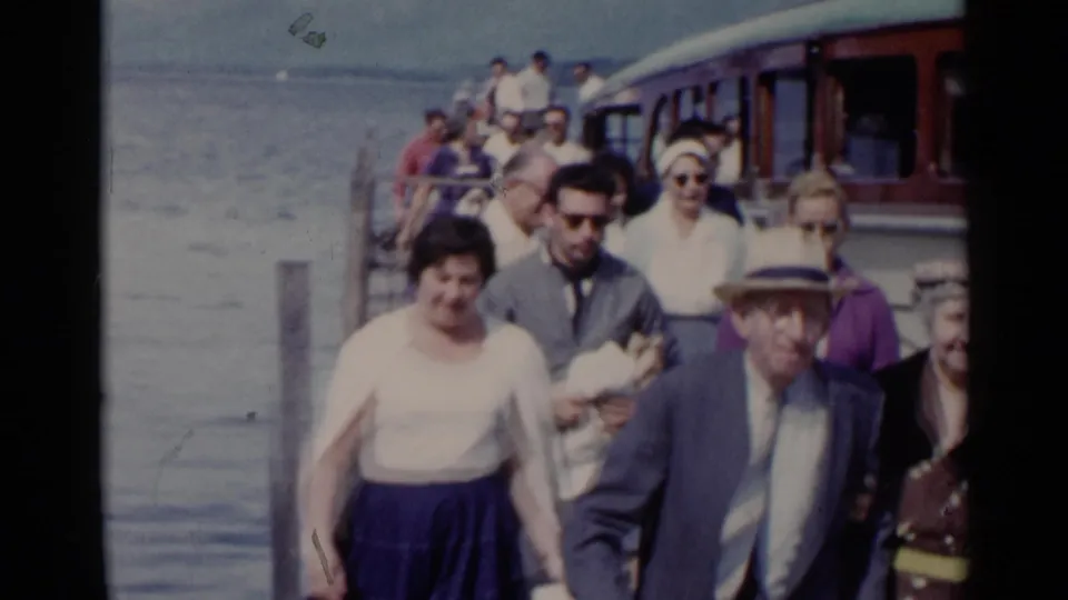 In Lake George, New York, 1955 home-movie footage, adults in mid-century clothing walk on a dock beside the lake.