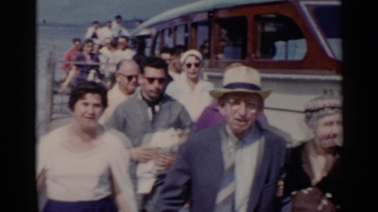 Passengers in 1950s attire walk toward the camera on a Lake George dock, with a white and brown motorboat behind them.