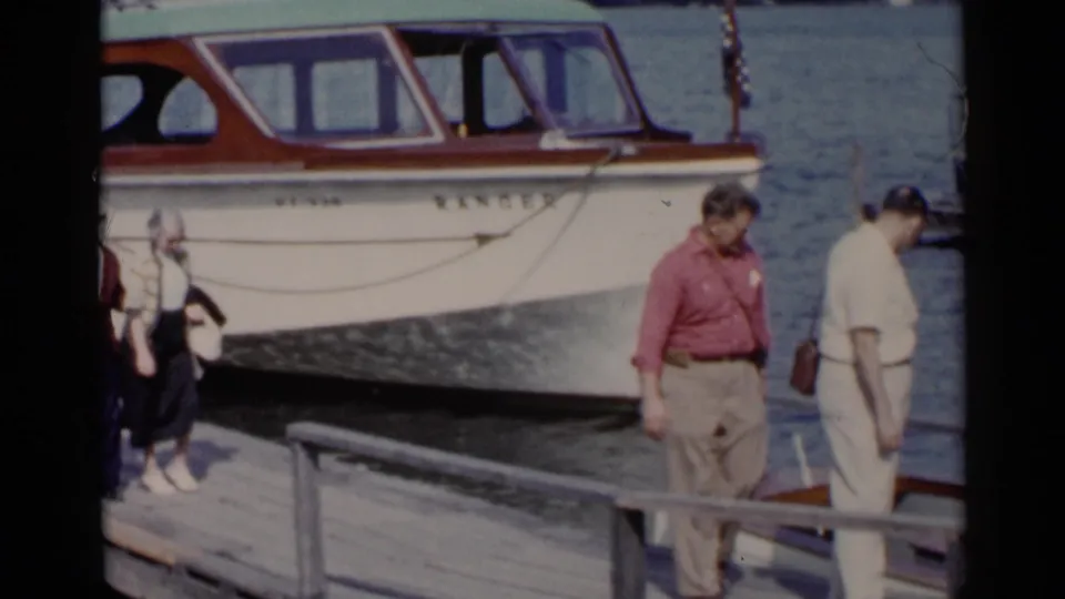 A white and brown boat marked RANGER sits by the dock as adults walk and pause near the water in Lake George.