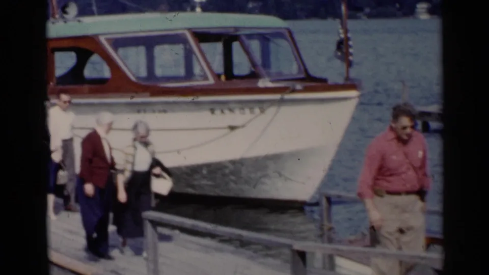 Adults in hats and cardigans walk along the Lake George dock beside the moored RANGER boat in 1955 home-movie footage.